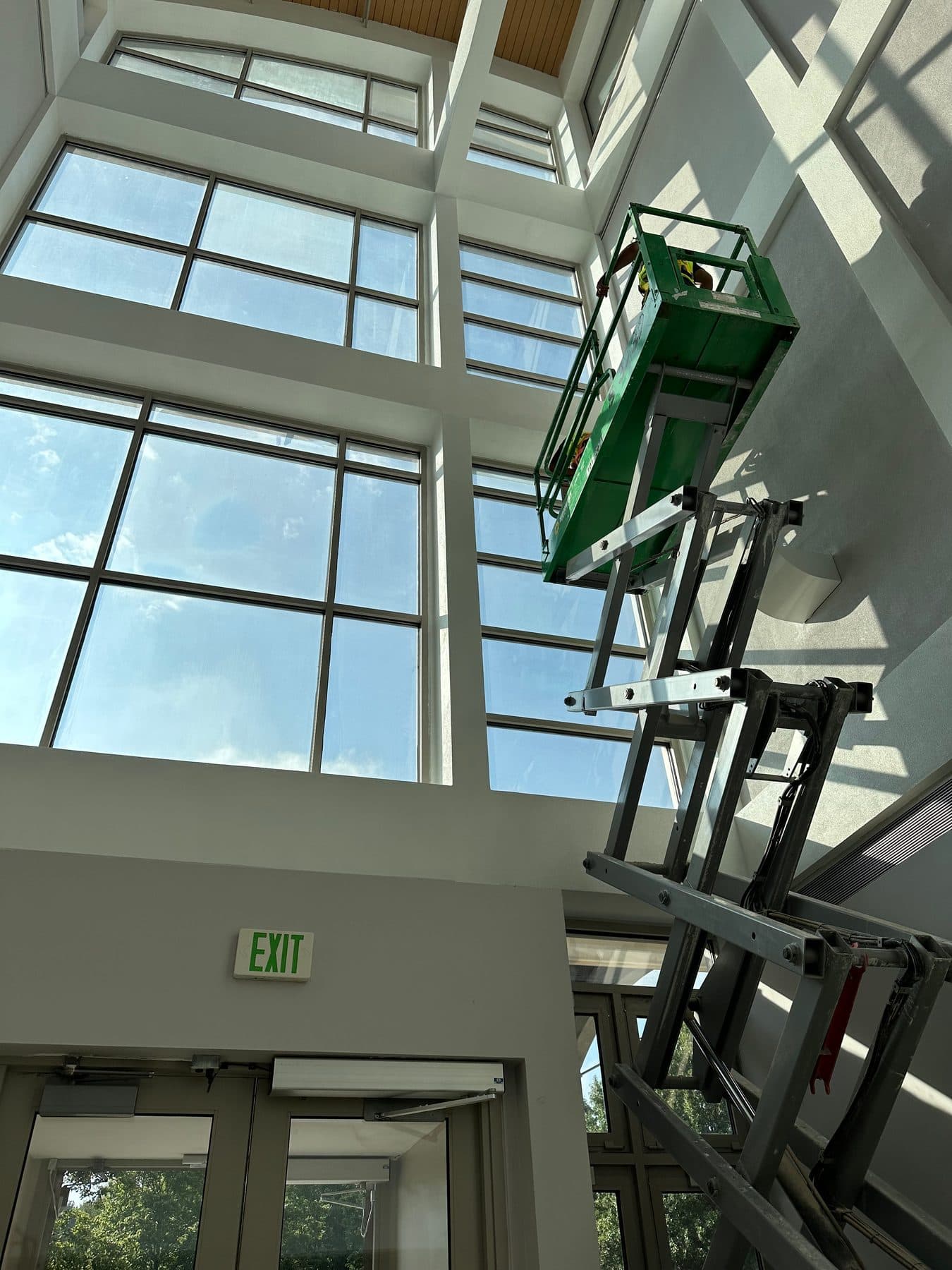 A High Five cleaner on a green lift, washing tall interior windows in a commercial atrium