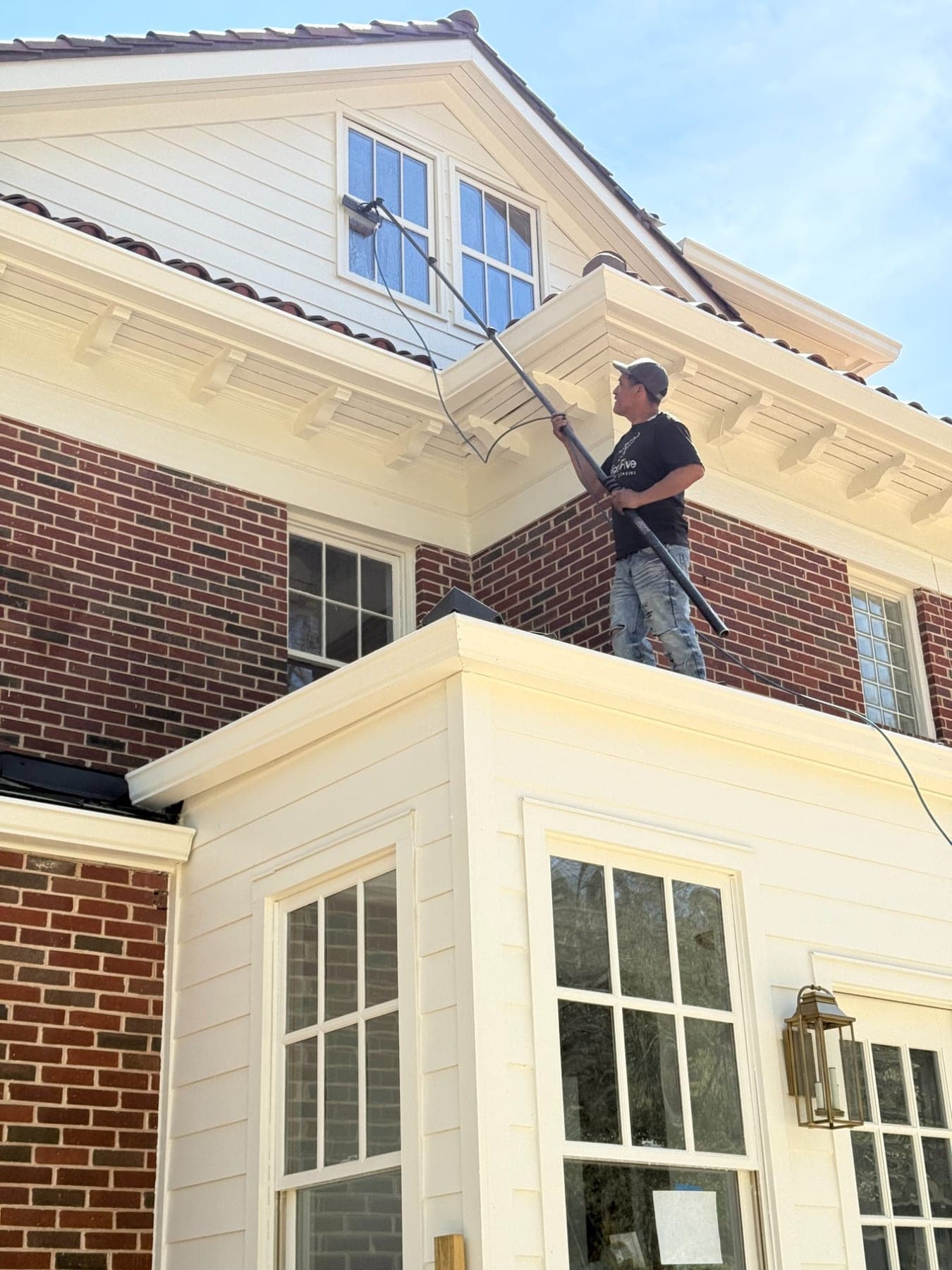 A cleaner standing on a porch roof, reaching a dormer window with a water-fed pole