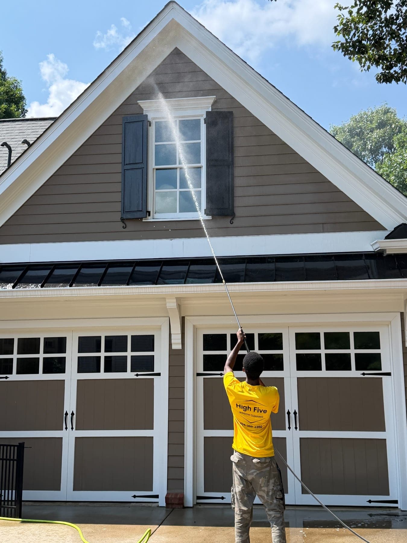 A High Five cleaner in a yellow shirt washing the siding above a garage